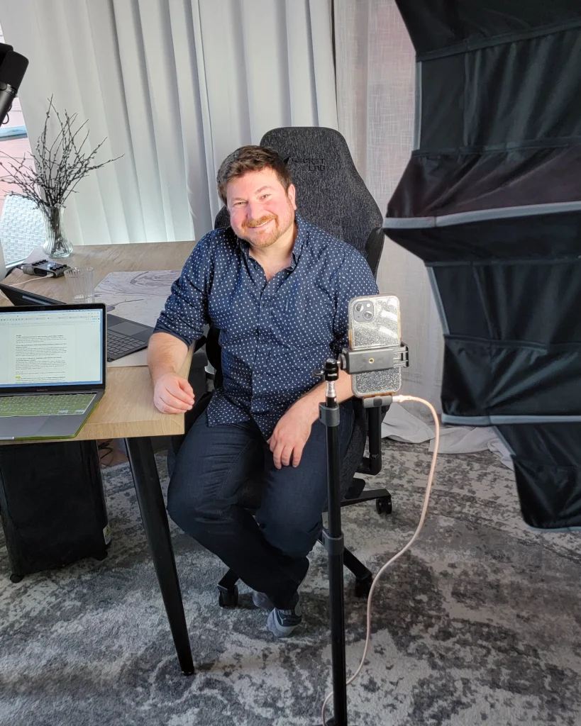 Craig sitting at a desk smiling at the camera. To his left is a laptop and microphone and to his right a light and cell phone on a stand. It's a behind the scenes look at filming for social media content creation.